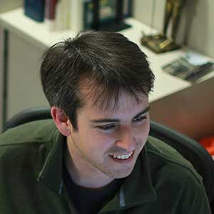 An overhead view of Ryan Marx, smiling while he works, sitting at his desk. The walls of his cubicle are covered in small posters and family photos. Ryan is wearing one of his favofite fleece zip-up jackets.